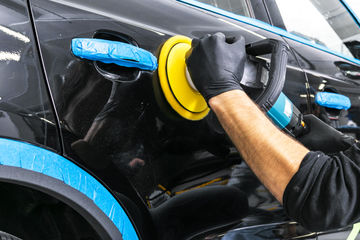 Gloved person polishing a black car door with a buffer in an auto detailing shop